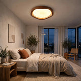 A modern bedroom interior with a bed dressed in neutral beige bedding, a wooden nightstand, potted plants, and a large oval ceiling light glowing above. Large windows with sheer curtains look out onto a dark evening sky.