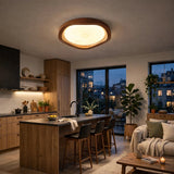 A modern oval ceiling light with a warm glowing white center and a natural wood frame, installed in a contemporary kitchen and living room setting.