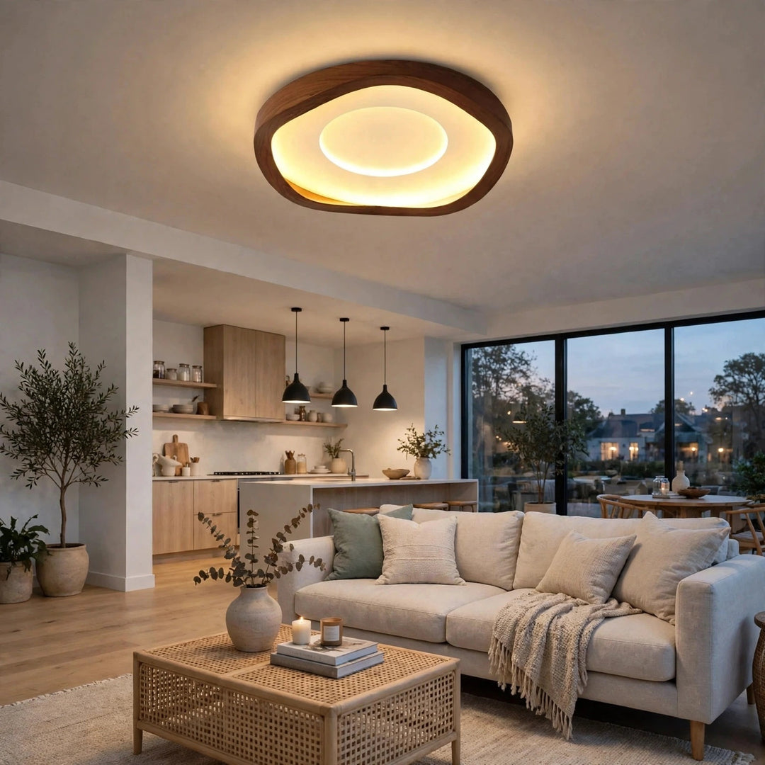 A modern living room featuring a large, organic-shaped ceiling light with a dark wood frame and warm glowing LED ring. The room is styled with a light beige sofa, a woven rattan coffee table, and a kitchen area in the background.
