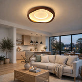 A modern living room featuring a large, organic-shaped ceiling light with a dark wood frame and warm glowing LED ring. The room is styled with a light beige sofa, a woven rattan coffee table, and a kitchen area in the background.