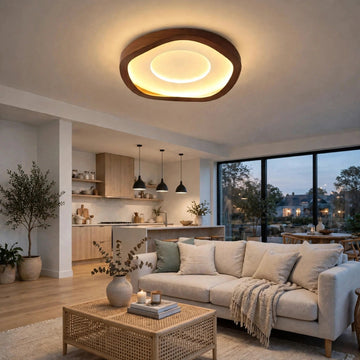 A modern living room featuring a large, organic-shaped ceiling light with a dark wood frame and warm glowing LED ring. The room is styled with a light beige sofa, a woven rattan coffee table, and a kitchen area in the background.