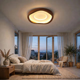 A modern oval ceiling light with a warm glowing LED ring and a dark wood frame, installed in a beautifully styled, minimalist bedroom with beige bedding, a rattan chair, and large windows overlooking a city skyline at dusk.