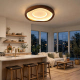 A modern, organic-shaped ceiling light fixture with a warm glowing LED ring and a dark wood frame, installed in a bright, contemporary loft apartment with a kitchen, bar stools, and a living room.