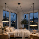 Two black pendant lights with large dome shades hang over a neatly made bed in a modern bedroom with large windows overlooking a city skyline at dusk.