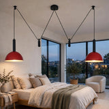 A modern bedroom interior with two red pendant lights hanging from black cords above a bed. Large floor-to-ceiling windows show a city view at dusk, and a white armchair and potted plant sit nearby.