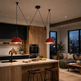 Two modern pendant lights with red dome shades and black cords hang over a kitchen island in a contemporary living space with wood cabinets, a sofa, and large windows.