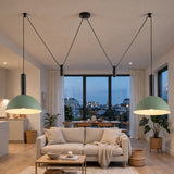 A modern living room with two large sage green pendant lights hanging from black cords above a beige sofa. The room has a minimalist aesthetic with light wood floors, a low wooden coffee table, and large windows showing a city view at dusk.