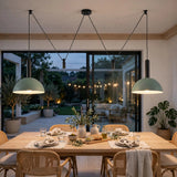 A modern dining room with a light wood table, woven chairs, and two large sage green pendant lights hanging over the table. Large black-framed glass doors open to a backyard patio at dusk, with plants and string lights visible outside.