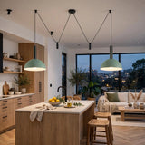 Two sage green pendant lights with black cords hang over a modern kitchen island in a bright, contemporary living space with wood cabinetry, a sofa, and large windows.