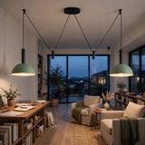 A modern living room with two pendant lights featuring sage green dome shades and black cord suspensions, hanging over a wooden desk and seating area with armchairs and a view of the city skyline at dusk.