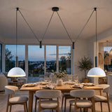 Modern dining room with a wooden dining table, upholstered chairs, and four black pendant lights with white dome shades hanging over the table. Large windows in the background show an evening city view.