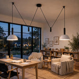 Two modern pendant lights with white dome shades and black cords hang over a cozy living room setup with a wooden desk, armchairs, and large windows showing an evening outdoor scene.