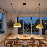 Two modern pendant lights with bright yellow dome shades hang over a wooden dining table set for dinner in a bright, contemporary dining room with large windows.