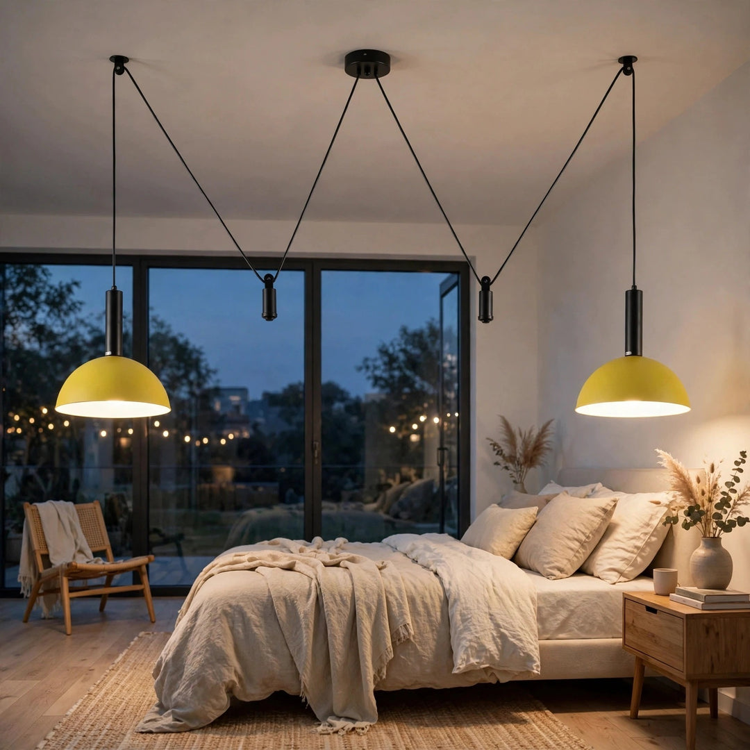 Two modern pendant lights with yellow dome shades and black cords hang above a bed in a styled bedroom. The room has a large window looking out onto a dusk scene with string lights, a wooden chair, and a bedside table.