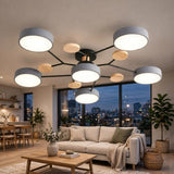 A modern multi-light ceiling fixture with six cylindrical white shades and light wood accents on a black frame, shown installed in a contemporary living room with a sofa, coffee table, and large windows overlooking a city skyline at dusk.