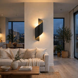 A modern spiral-shaped wall sconce with a matte black finish, mounted on a beige wall in a contemporary living room. The warm light emanates from the center of the spiral, illuminating a beige sofa, wooden coffee table, and large windows in the background.
