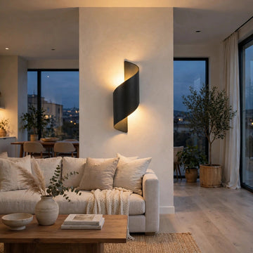 A modern spiral-shaped wall sconce with a matte black finish, mounted on a beige wall in a contemporary living room. The warm light emanates from the center of the spiral, illuminating a beige sofa, wooden coffee table, and large windows in the background.