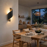 A modern black spiral-shaped wall sconce mounted on a warm off-white wall in a beautifully styled dining room with a wooden table, woven chairs, and a large window view.