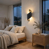 A modern black spiral-shaped wall sconce mounted on a beige wall in a styled bedroom, casting a warm light. The room features a wooden bed with neutral linens, a rattan nightstand with a vase of branches and an open book, and large windows looking out onto a dusk scene.