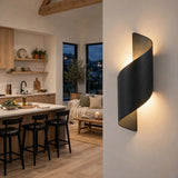 A modern spiral-shaped wall sconce in a dark matte finish, mounted on a warm off-white wall and casting a soft glow. The background shows a contemporary kitchen and living room interior with light wood floors, a white island, and black bar stools.