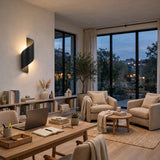 A modern spiral black wall sconce mounted on a warm beige wall in a stylishly decorated living room with large windows, armchairs, a wooden desk, and a view of the city at dusk.
