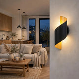 A modern spiral-shaped wall sconce with a matte black body and a warm yellow-gold inner panel, mounted on a textured wall in a contemporary living room setting.