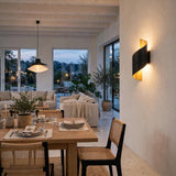 A modern black wall sconce with a warm gold interior glow, mounted on a textured off-white wall in a beautifully styled dining room with a wooden table, woven chairs, and large windows overlooking a patio.
