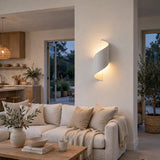 A modern spiral-shaped wall sconce with a warm white shade mounted on a beige wall in a beautifully styled living room with a cream sofa and wooden coffee table.