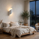 A modern spiral wall sconce with a white curved body and warm glowing light, mounted on a textured beige plaster wall above a minimalist bed in a cozy, neutral-toned bedroom.