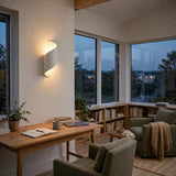 A modern spiral-shaped wall sconce with a white finish, glowing with warm light on a beige wall in a cozy, contemporary home office and reading nook with large windows, a wooden desk, and green armchairs.