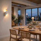 A modern wall-mounted sconce with a white sculptural shell and warm yellow light glowing from within, mounted on a beige wall in a contemporary dining room setting.