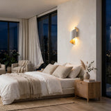 A modern wall-mounted sconce with a white rectangular body and warm yellow-gold interior, casting light onto a textured beige wall in a styled bedroom setting.