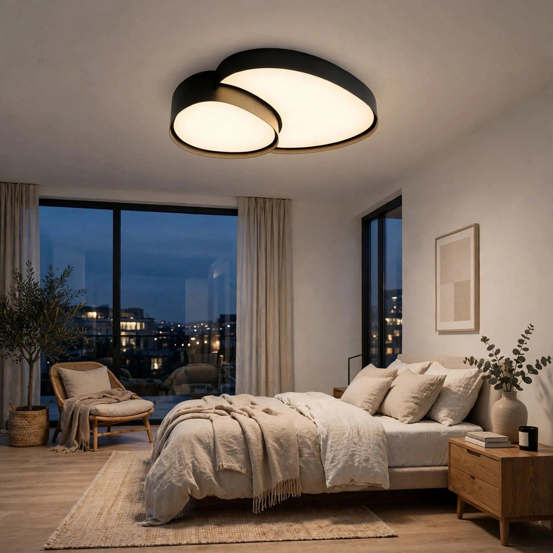 Modern two-ring oval ceiling light fixture with black frames and warm white glowing bulbs, installed in a contemporary bedroom with a bed, nightstand, and large windows overlooking a city skyline at dusk.