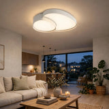 White flush-mount ceiling light with overlapping rounded illuminated panels above a neutral living room with a cream sofa, wooden coffee table, and plants.
