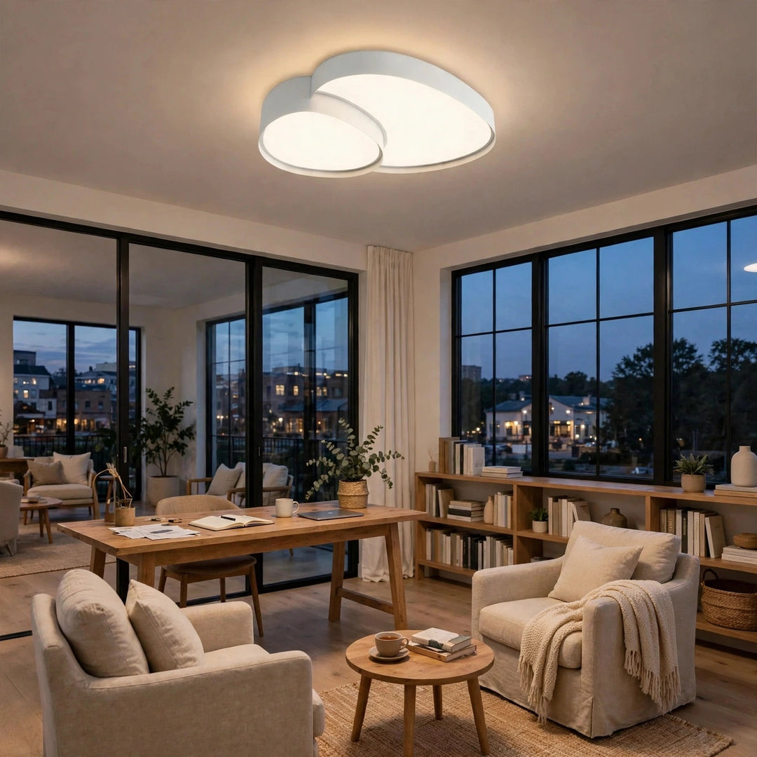 A modern cloud-shaped ceiling light fixture with two overlapping rounded white diffusers, glowing with warm light in a contemporary living room with beige armchairs, a wooden desk, and large black-framed windows.