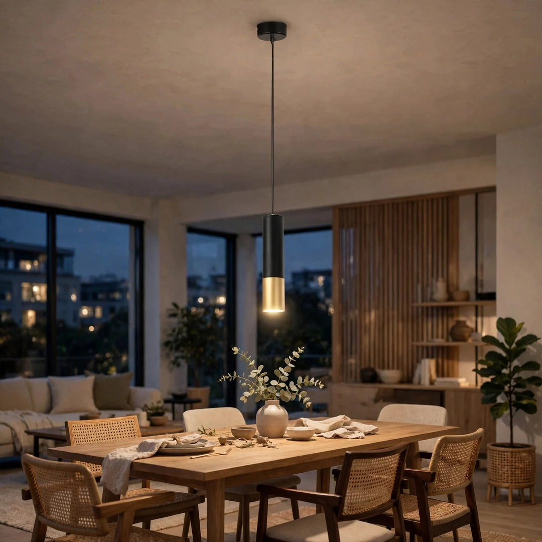 A modern cylindrical pendant light with a matte black body and a brushed gold metallic bottom, hanging over a wooden dining table in a warm, contemporary dining room.