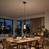 A modern cylindrical pendant light with a matte black body and a brushed gold metallic bottom, hanging over a wooden dining table in a warm, contemporary dining room.