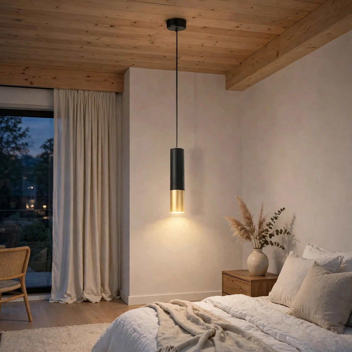 A modern cylindrical pendant light with a matte black upper body and a brushed gold lower shade, hanging in a cozy, minimally styled bedroom with a wooden ceiling and beige walls.