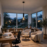 A black cylindrical pendant light with a glowing gold-colored tip hangs in a home office with beige armchairs and large windows.