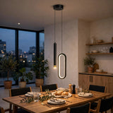 Modern two-light pendant dining fixture in a real dining room setting, with one slim cylindrical black lamp and one elongated oval black frame lamp glowing with warm light, above a wooden dining table set for dinner.