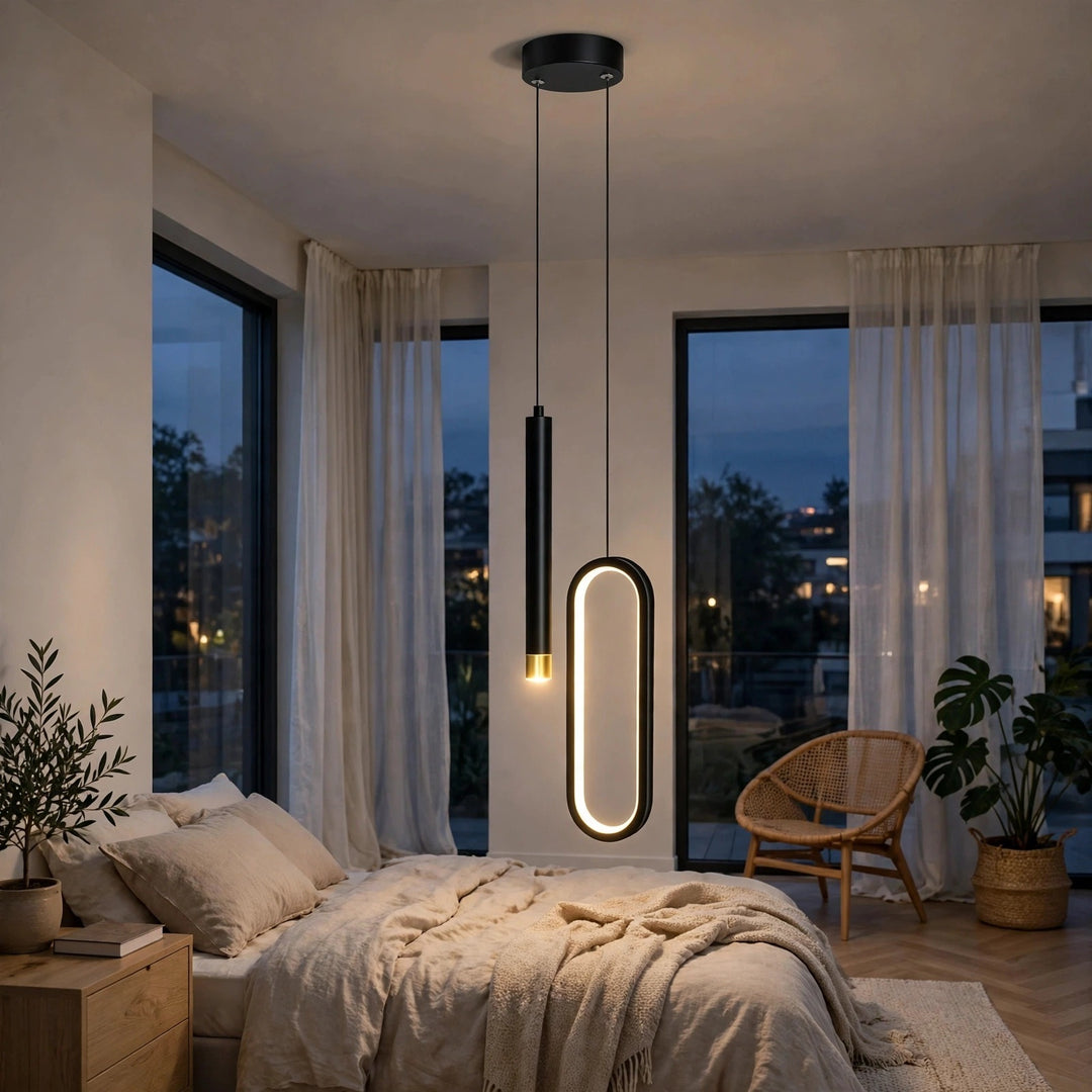 A modern two-light pendant ceiling fixture hangs over a bed in a softly lit bedroom. One lamp is a slim black cylindrical rod with a small gold-toned accent near the bottom, and the other is a rounded vertical oval with a warm white illuminated edge and black frame.