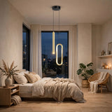 A modern bedroom interior with a glowing linear pendant light hanging above a bed. The room has warm neutral walls, beige bedding, wood flooring, a window at dusk, plants, shelves, and a wicker chair with a throw blanket.