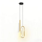 Modern pendant light fixture with a black circular ceiling mount, two slim black suspension cords, one gold cylindrical shade, and one elongated gold oval frame, shown against a plain white wall with warm light glow.