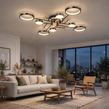 Modern multi-light ceiling fixture with circular LED rings and a brushed brass center, installed in a contemporary living room with a sofa, coffee table, and large windows overlooking a city skyline at dusk.