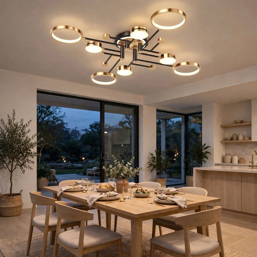 Gold and black ceiling light with circular illuminated discs above a wooden dining table in an open kitchen at dusk.