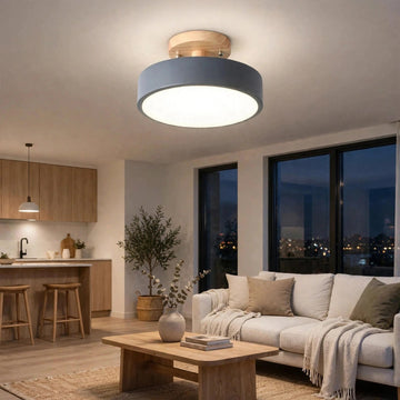 A modern flush-mount ceiling light with a circular blue-gray shade and a light wood ceiling mount, installed in a bright, contemporary living room with a sofa, coffee table, and kitchen in the background.