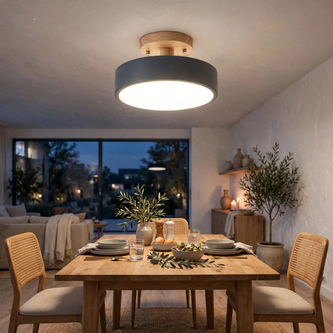 A modern semi-flush mount ceiling light with a matte dark gray cylindrical shade and a light wood top, glowing warmly above a wooden dining table in a styled home interior.