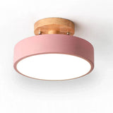 A modern flush-mount ceiling light with a matte pink cylindrical shade, a natural wood ceiling mount, and a glowing white diffuser, shown against a plain white background.