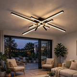 Black crossed linear-bar ceiling light glows in a living room with rattan armchairs and glass doors opening to a patio at dusk.