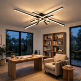 Modern ceiling light fixture with multiple linear LED tubes in a black and brushed silver metallic finish, mounted on the ceiling of a contemporary home office with a wooden desk, armchair, and large windows.